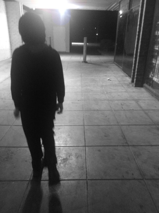 When my son was little and he would be crying and his dad would be shitty I’d go for walks at 3am around the inner city. Even though it was dark it was safe – safer than being at home. A black and white image of an empty concrete undercover brightly lit sidewalk. On the left side of the image, the silhouetted figure of a young person appears to be walking toward the camera. We cannot make out any features.