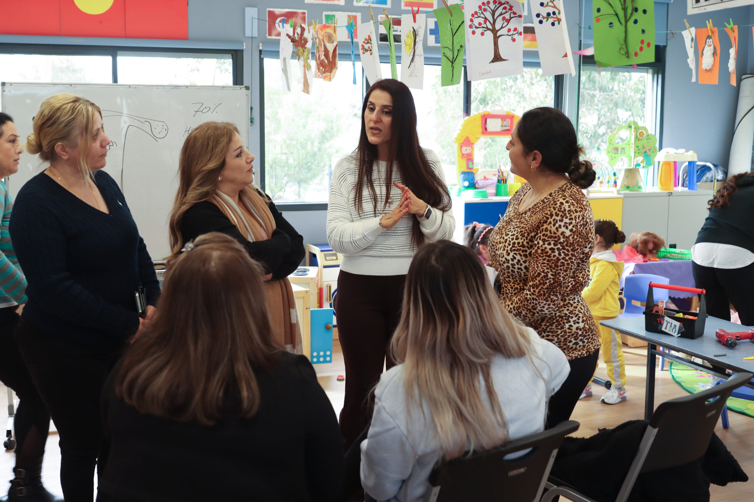 A woman speaks to a diverse group of women in a classroom setting. They are standing and listening to her intently