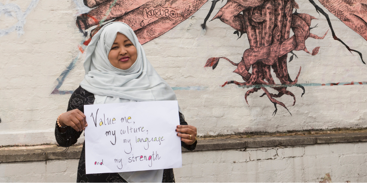 A woman with a beautiful smile in a light grey hijab holding a sign that reads 'Value me, my culture, my language and my strength'