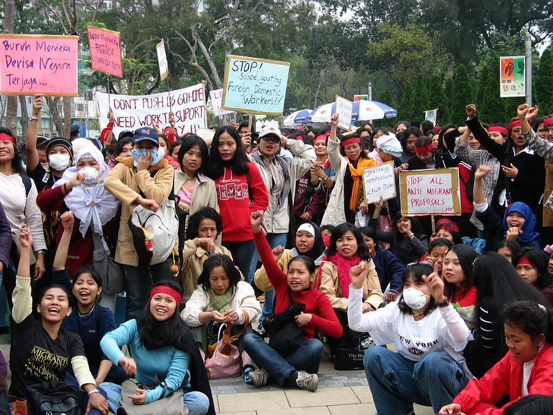 A large group of women sitting and standing outside together from the Migrant Centre Hong Kong holding placards