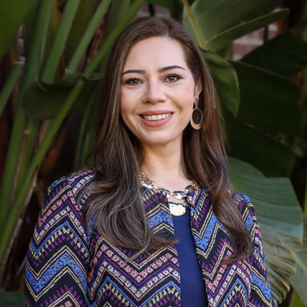 Eman Al-Dasuqi is looking at the camera and smiling. She is wearing a a patterned blouse and is photographed in an outdoor natural setting.
