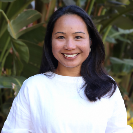 Farhana Kuthupdeen is looking at the camera and smiling. She is wearing a white shirt and is photographed in an outdoor natural setting.