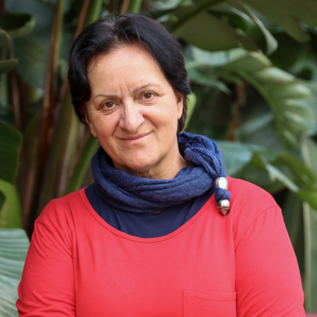 Felicia is smiling at the camera against a natural background. She is wearing a navy turtleneck with a red jumper over the top.