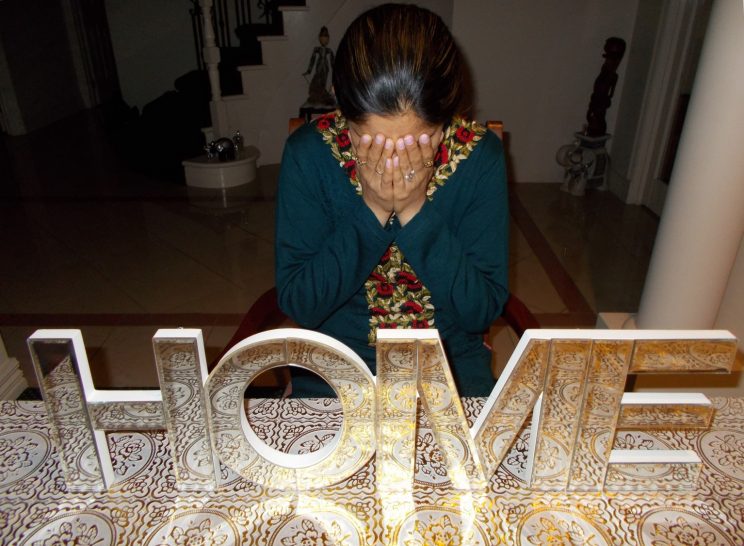 A woman sits in a rich looking home, leaning forward with her hands covering her face. She is wearing a teal top or dress, which is richly embroidered down the front with red, gold and olive flowers. Directly in front of the woman, large wooden mirrored letters that spell the word HOME have been arranged to fill the bottom of the image. The letters sit on a bench tiled with an ornate gold floral print, which is reflected in the letters, and makes them appear gold. Behind the woman we can see a staircase and decorations.