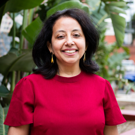 An image of Monisha from MCWH smiling at the camera in a natural setting. She is wearing a red top.