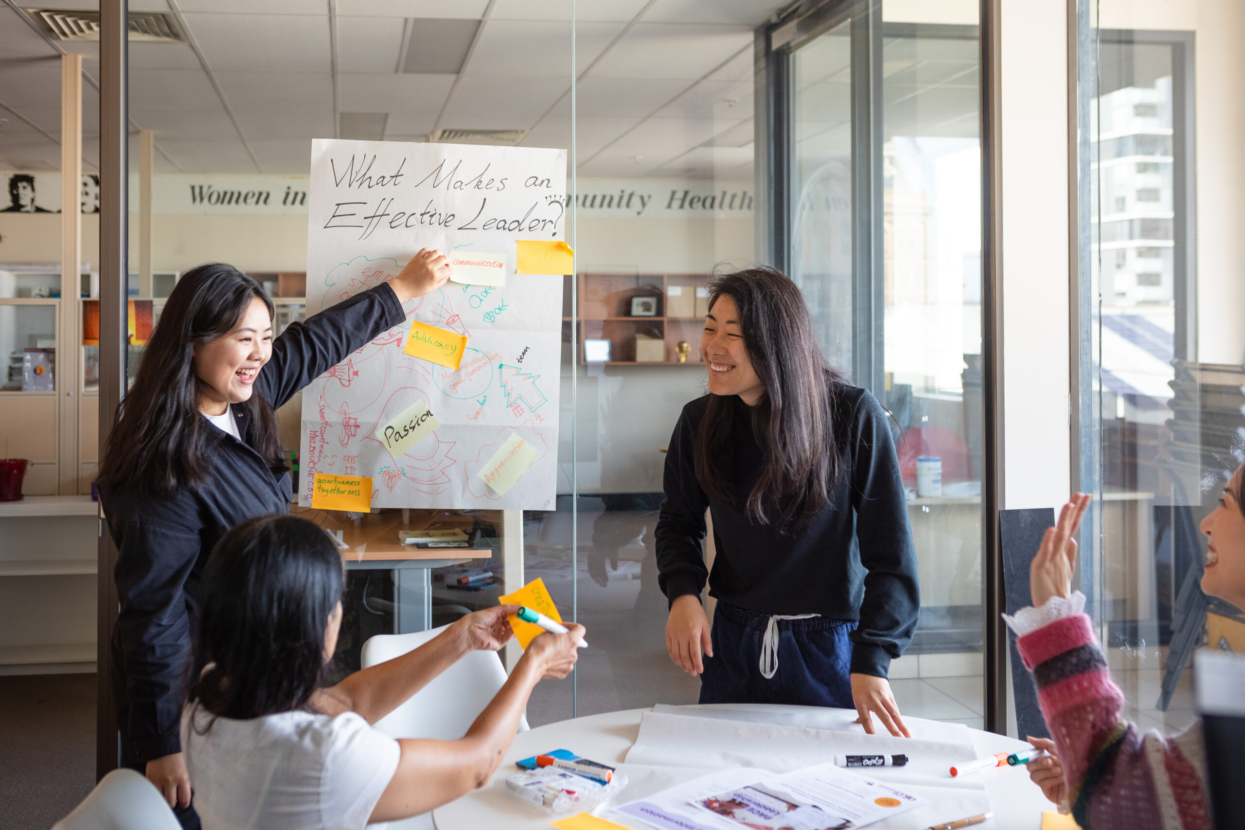 Three women sit at a conference table taking notes on a window with sticky noted about what makes an effective leader
