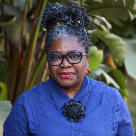 Tamara Kwarteng is smiling slightly at the camera wearing a blue shirt against a green outdoor background.