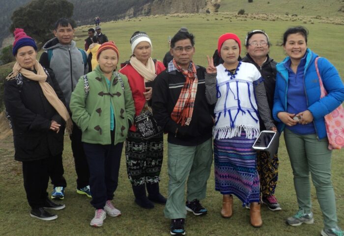 This is a photograph of members of the Karen community in Hobart. We want to send a message that in the Karen community we want to be free from violence. We want to be as happy as we are in this picture. A candid image of eight Karen community members posing for a different picture, looking just out of frame to the right. The group includes men and women of range f ages, some smiling and others not. They are all wearing warm clothing and many have beanies. Behind them, a large grassy field stretches towards a mountainous looking background. We can see other people around in the background, enjoying the view. One woman in the front is making a peace sign for the photograph.
