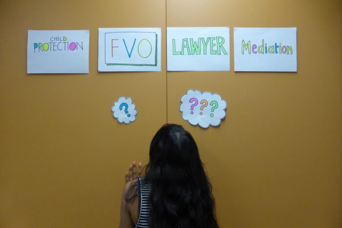 Women who are going through the system find it so overwhelming. Even people who grew up here would find it confusing but for us, it’s so hard. Even when they get to family court they often have no idea what’s happening. Mediation. What’s mediation? Lawyer. What’s lawyer? They think everything that is offered to them must be to help them so they accept it. But they don’t necessarily understand it. A figure with long black hair stands with their back to the camera, looking up at a yellow wall. There are four pieces of paper on the wall. The first reads ‘Child Protection’, the second ‘FVO’, the third ‘lawyer’ and the fourth ‘mediation. Two pieces of paper below with question marks sit just above the head of the figure, as if they were cartoon thought bubbles.