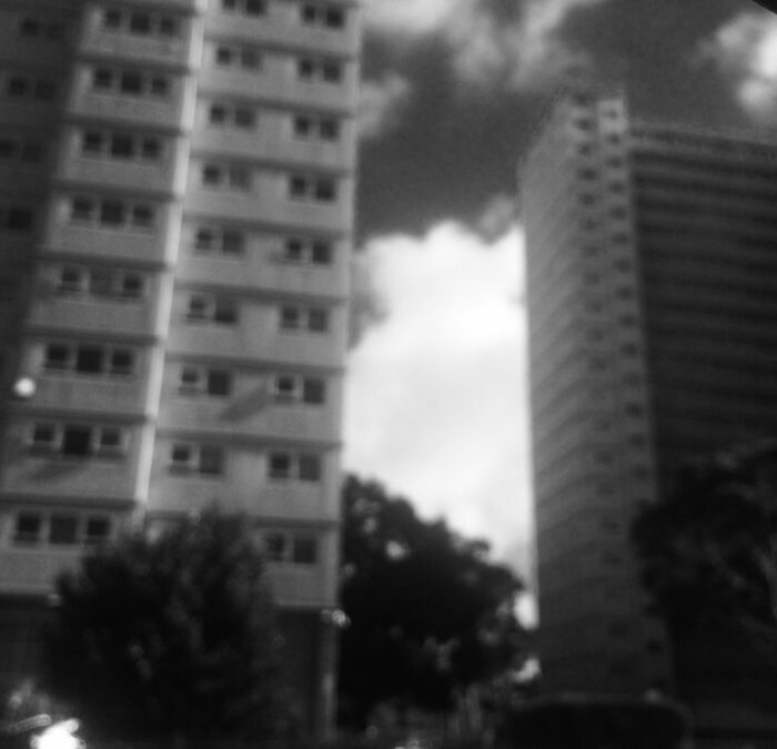 When I went back to take photos I was surprised by how afraid I was and I couldn’t stop the car, so the image is blurred. It’s hard realising that it has been so many years, but the feelings are still there. A black and white image of a public housing high rise on a cloudy day. The image is blurry and the angle of the image seems slightly tilted.