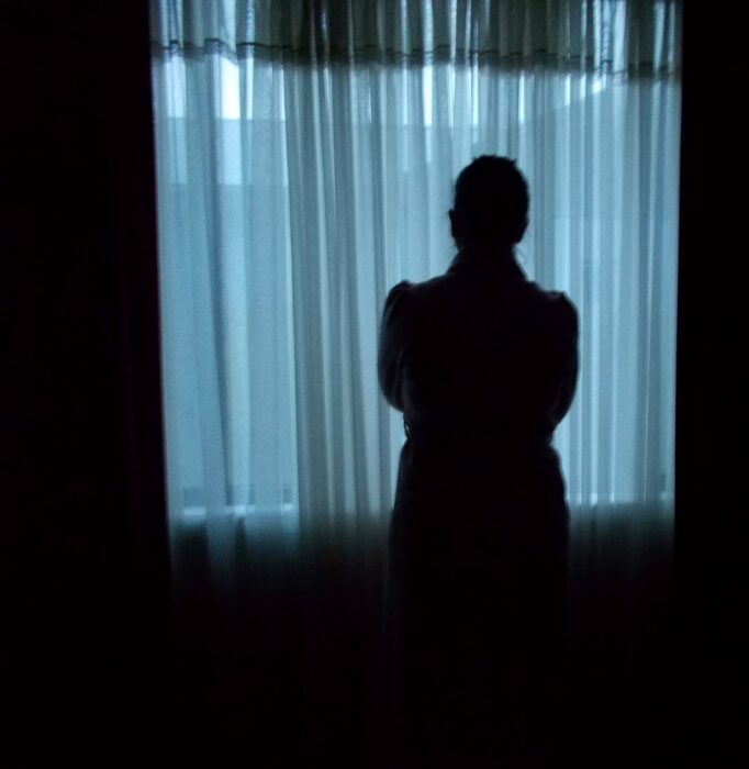 When you do not have security, it is not easy to trust. You withdraw and your world reduces considerably. What Services and the Police do not take into consideration is how this impacts on the individual and exposes them to vulnerability causing anxiety and fear.
Imagine you are the person in this image. You are looking at the world behind a sheer curtain. Can you see clearly or is the curtain blurring your vision? Add to this a fear of the outside world that has been imposed on you by an abuser, who controls you with phrases like, ‘Where do you think you are going to go?’ or ‘Who do you think is going to listen to you?’ Now be honest and don’t say you would get yourself out of there because homelessness is a reality when you have no money, no contacts, no food security, your self-esteem has been totally crushed and you cannot speak the English language. Who would YOU trust? A silhouetted figure stands in a dark room, in front of a window with a drawn muslin curtain. The light that comes through the curtain casts a cool blue light. The figure appears to have their arms crossed in front of their body.