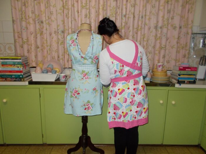 My mother before me had no voice. In her generation women were told, “it is a sin to speak against your husband”. As a consequence, women took their secrets to their graves exposing another generation of women to abuse. A woman stands with her back to the camera and her head resting on the shoulder of a dressmaker mannequin, positioned in front of a kitchen sink. The woman and the mannequin are both in bright floral aprons and the kitchen sink and benches are piled with pastel crockery and stacks of brightly coloured cook books. The soft pink floral curtain above the kitchen sink is fully closed. The kitchen cupboards below the sink and benches are a bright lime green. In the far right corner of the kitchen bench, is a knife block and other hanging cooking utensils.