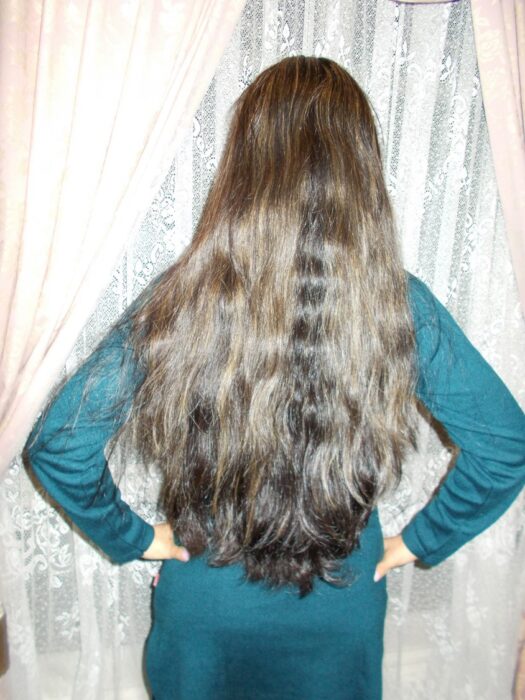 He was always threatening to cut off my hair. Other people complimented me. But he always complained and put me down about it. I started seeing myself the way he wanted me to see myself. But I also really was clinging to my hair. I took this image before I cut my hair. I never wanted to sacrifice the length. Later on, once I had left, I decided to cut my hair for myself. When I cut the hair, when I let go, I felt better. I had to let it go for myself. A woman stands with her back to the camera, in front of a window with lace curtains. She is showing the viewer her long dark hair, which spills down her back to below her waist. Her hair has golden brown highlights. She has her hands on her hips and she is wearing a long sleeved teal top or dress.
