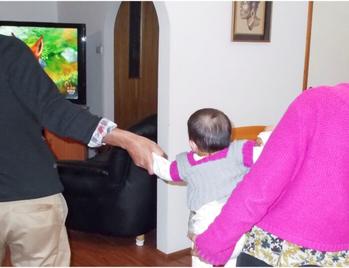 What I want to show with this photograph is that children are the victims of family violence. In a suburban loungeroom, we see the back of an infant with soft dark hair, in a onesie and knitted cardigan, being pulled in opposite directions by two figures, both mostly out of view. We can see one figure is wearing a bright pink cardigan and is moving out of the frame towards the right, while holding the infant. The other figure is wearing plain clothes and is grasping the arm of the infant and moving in the other direction, towards another room in which we can see a large television playing a kid's animation.