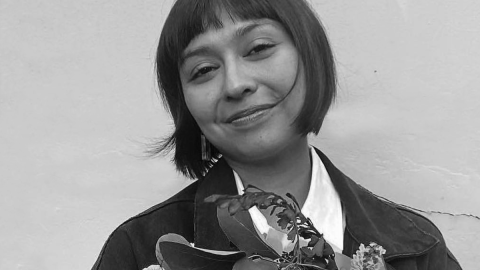 A close up photograph of a young woman with dark hair in a bob with bangs, smiling kindly and holding a bouquest of native Australian flowers. She is standing against a plain wall outside in natural light.